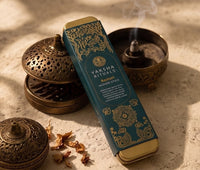 A Yaksha Rituals Kautilic incense stick box in deep teal with gold mandala designs, styled on a textured beige stone surface with brass incense burners and dried petals.