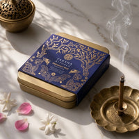 A square blue and gold tin of Yaksha Rituals Mahashaya Murti placed on a light marble surface, styled with a brass incense burner, a smoking incense stick holder, and flower petals.