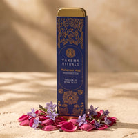 A tall blue box of Yaksha Rituals Maharani Mist Incense Stick with gold detailing and a gold cap, standing upright on a sandy surface surrounded by pink petals and small purple flowers under warm sunlight.