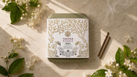 A square tin of Yansha Rituals Magna Sacra incense sticks is styled in a flat-lay on a light marble surface with white flowers, green leaves, and two wooden incense sticks.