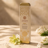 A Yaksha Rituals Mogra Incense Stick in a tall rectangular box with gold-toned metal ends, standing on a sandy surface with white flowers and soft sunlight in the background.