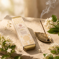 A Yaksha Rituals Muga Incense Stick in a cream-colored tin box with gold illustrations, styled on a stone slab with white flowers, a brass incense holder, and rising smoke.